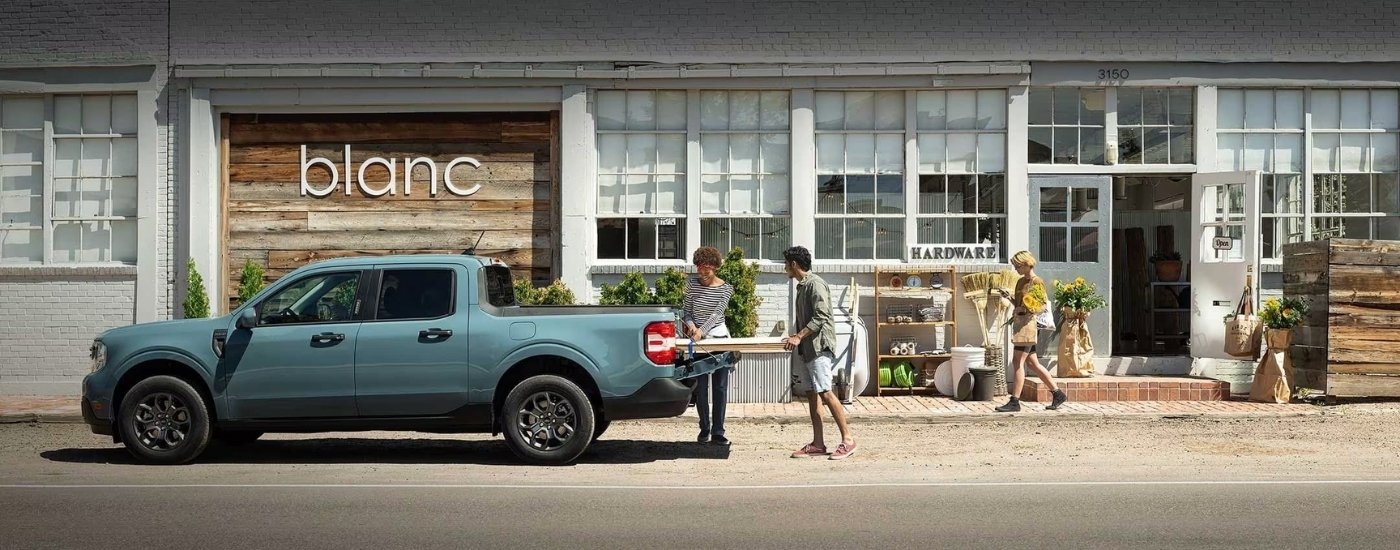 A blue 2023 Ford Maverick XLT is shown from the side in front of a shop.