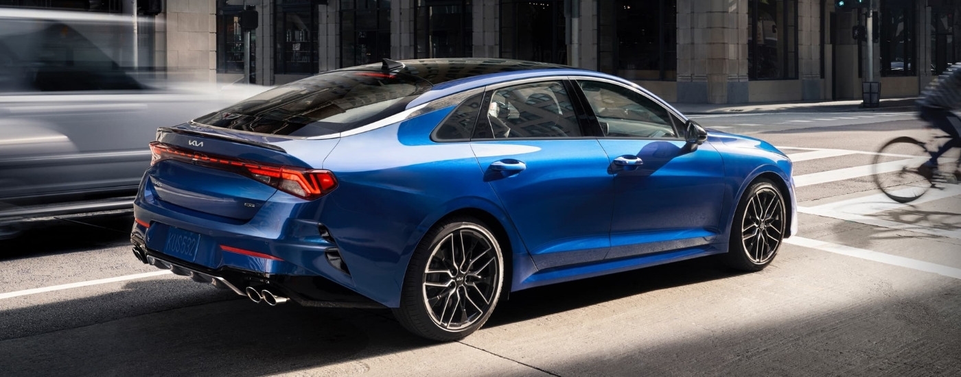 Rear view of a blue 2025 Kia K5 GT stopped a city intersection.