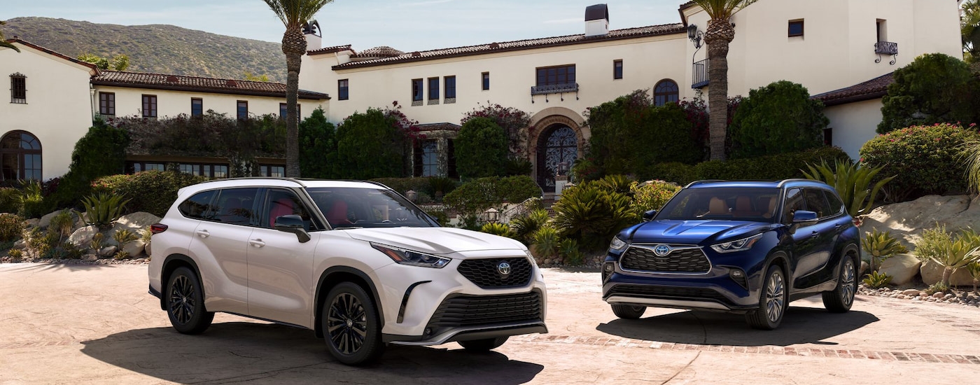 A white 2024 Toyota Highlander XSE and blue Hybrid Platinum parked in front of a large home.