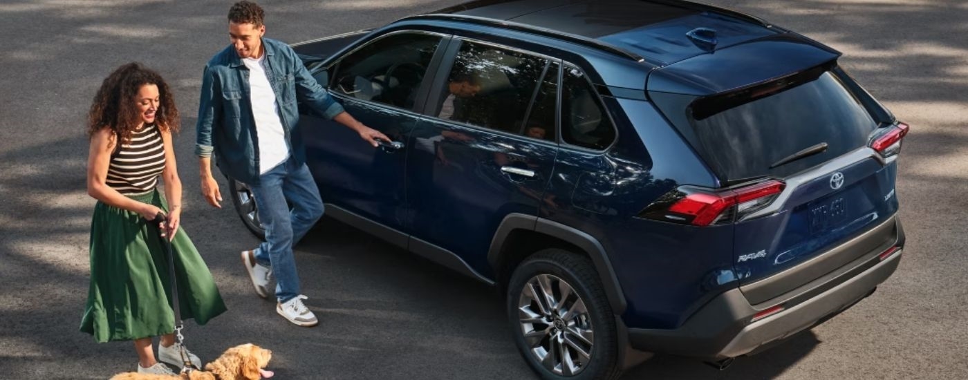 Two people and a dog are walking beside a parked blue 2025 Toyota RAV4.