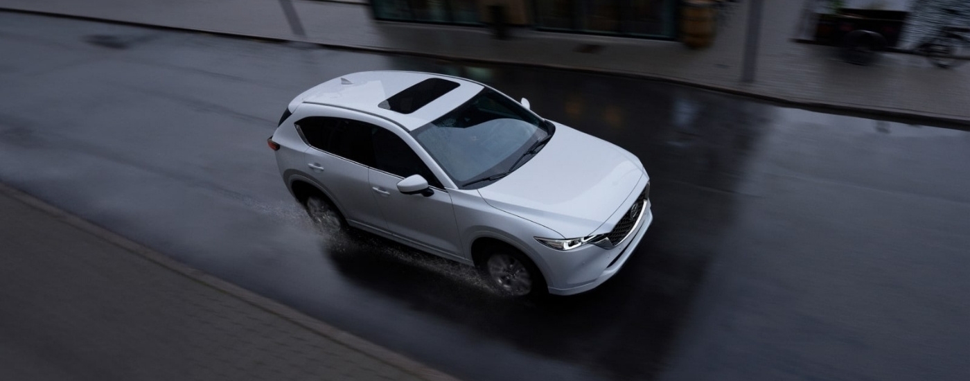 A white 2023 used Mazda CX-5 for sale is shown driving on wet pavement.