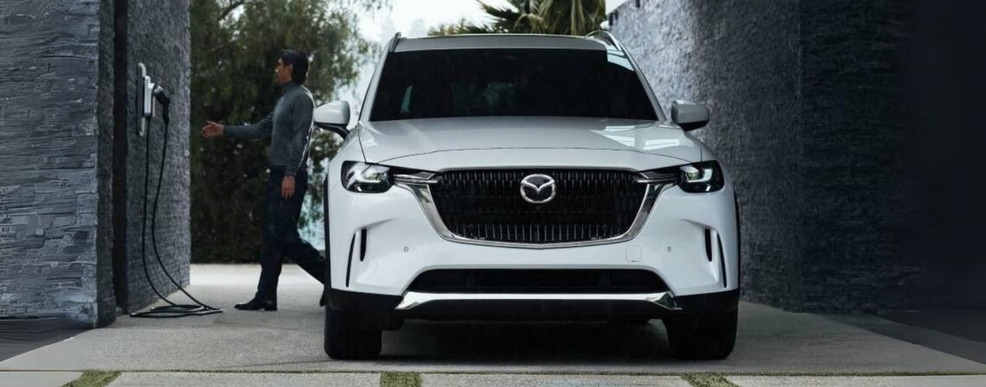 A white 2025 Mazda CX-90 PHEV is on a driveway near a charger.