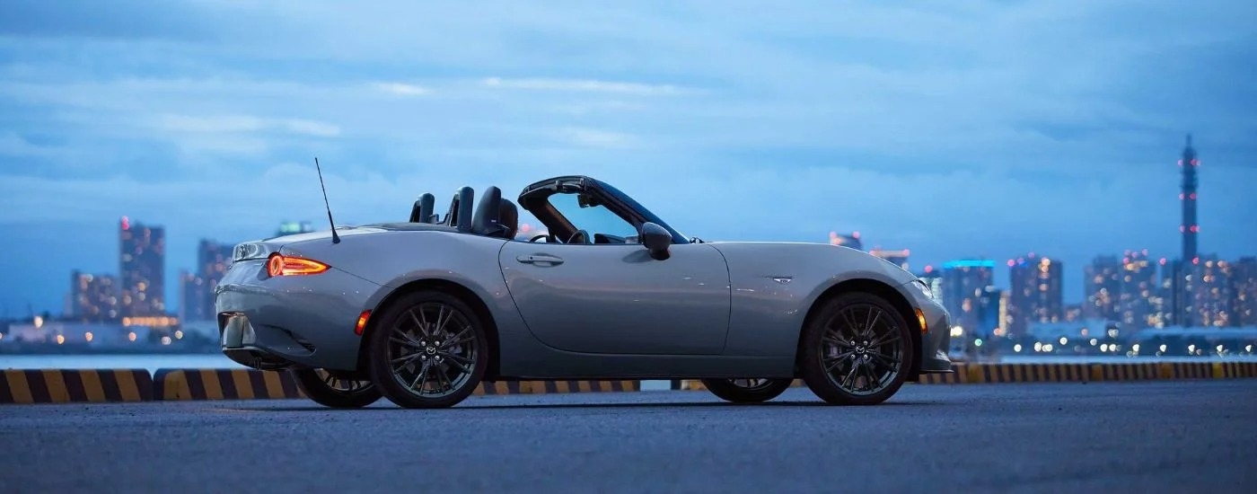 Side view of a light blue 2024 Mazda MX-5 Miata with the top down and a city skyline in the distance.