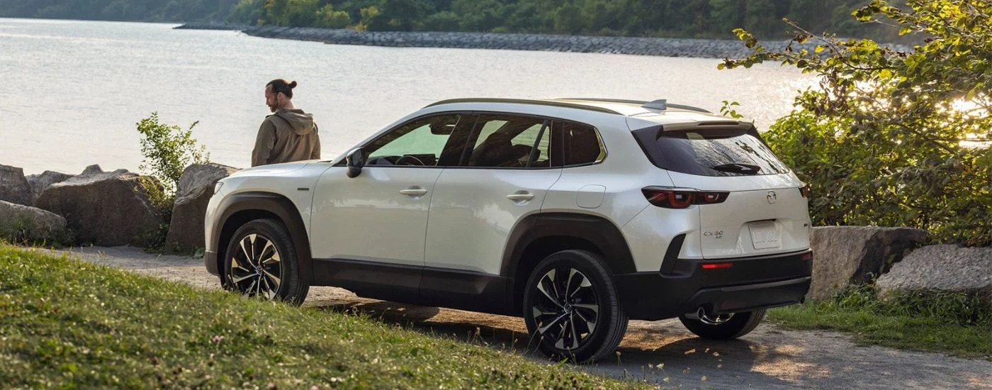 A man leaning up against a white 2025 Mazda CX-50 parked near a body of water.