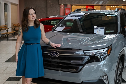 Woman posing with a new 2025 Subaru Forester in River Rock and the Johnstown Galleria Mall