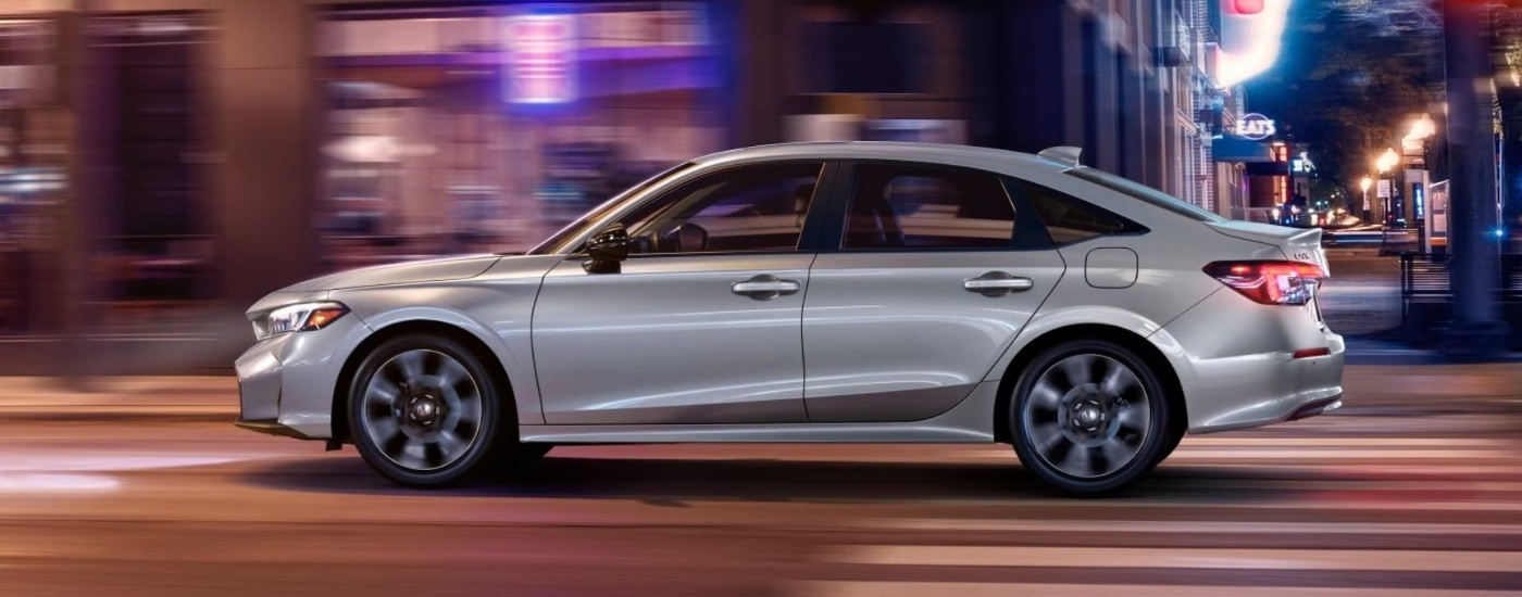 A silver 2025 Honda Civic Hybrid Sport Touring is driving to a Honda dealer.