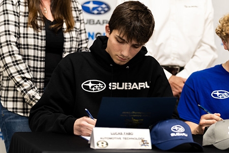Young man wearing black Subaru hoodie, sitting at a table, is signing a document