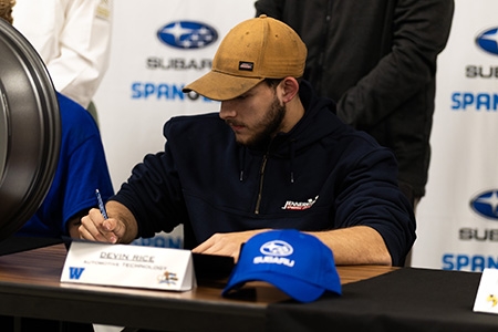 Student technician in hat is sitting at a table signing a contract with intent to work with Spangler Subaru