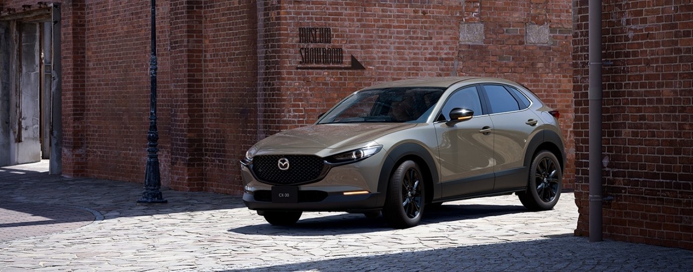 A tan 2025 Mazda CX-30 for sale near Kendall driving past a brick building.