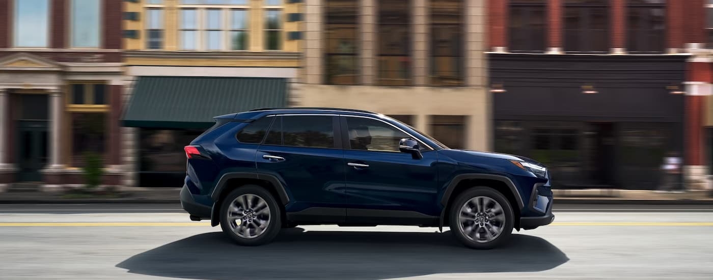 Side of a blue 2025 Toyota RAV4 Hybrid for sale near Rochelle driving on a city street
