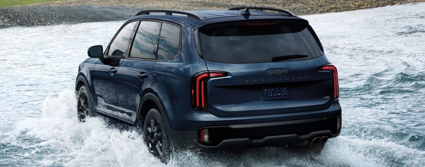Rear of a dark blue 2026 Kia Telluride driving over a shallow river.