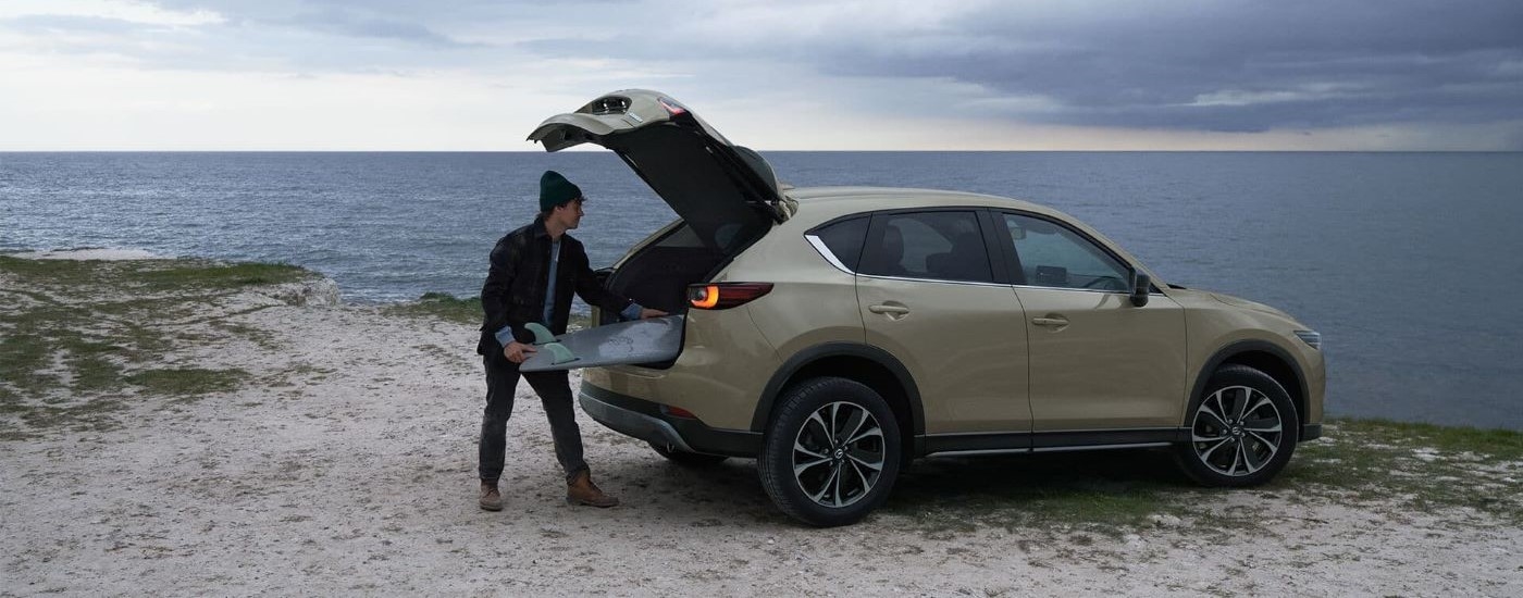 Person opening the hatch of a brown 2025 Mazda CX-5 parked on a coast.