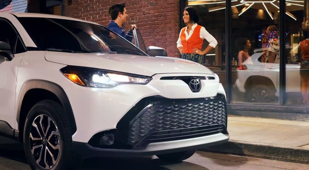 Close-up on the headlights and hood of a white 2026 Toyota Corolla Cross for sale near White Plains parked near a curb.