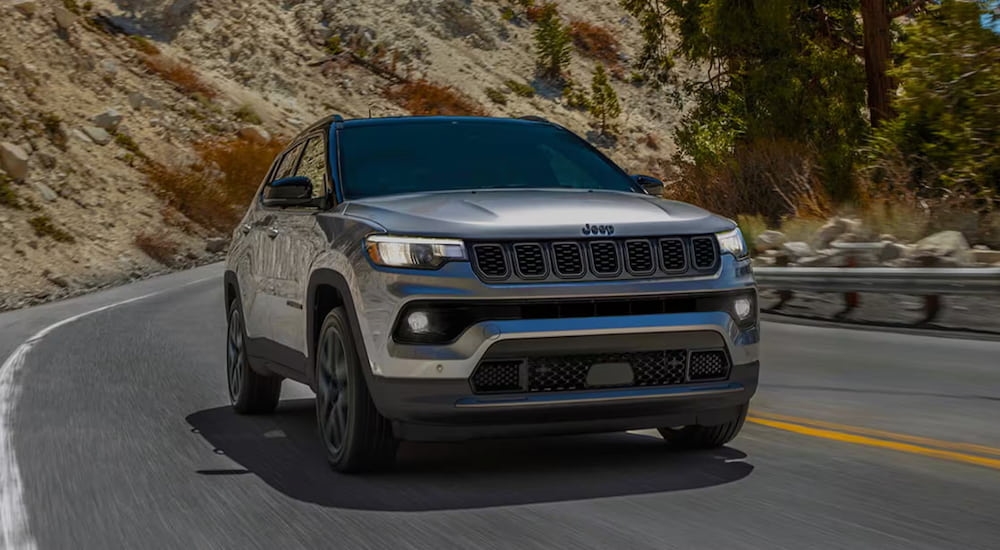 Grey 2025 Jeep Compass driving around a bend after visiting a car dealership.