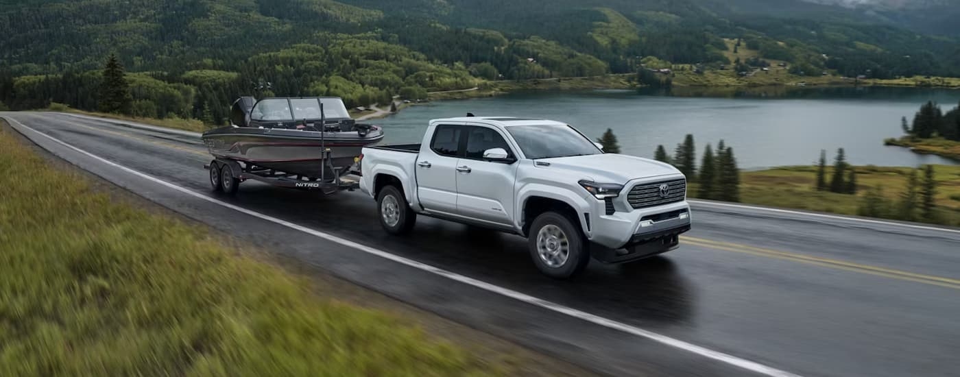 White 2026 Toyota Tacoma towing a boat on a road near a lake