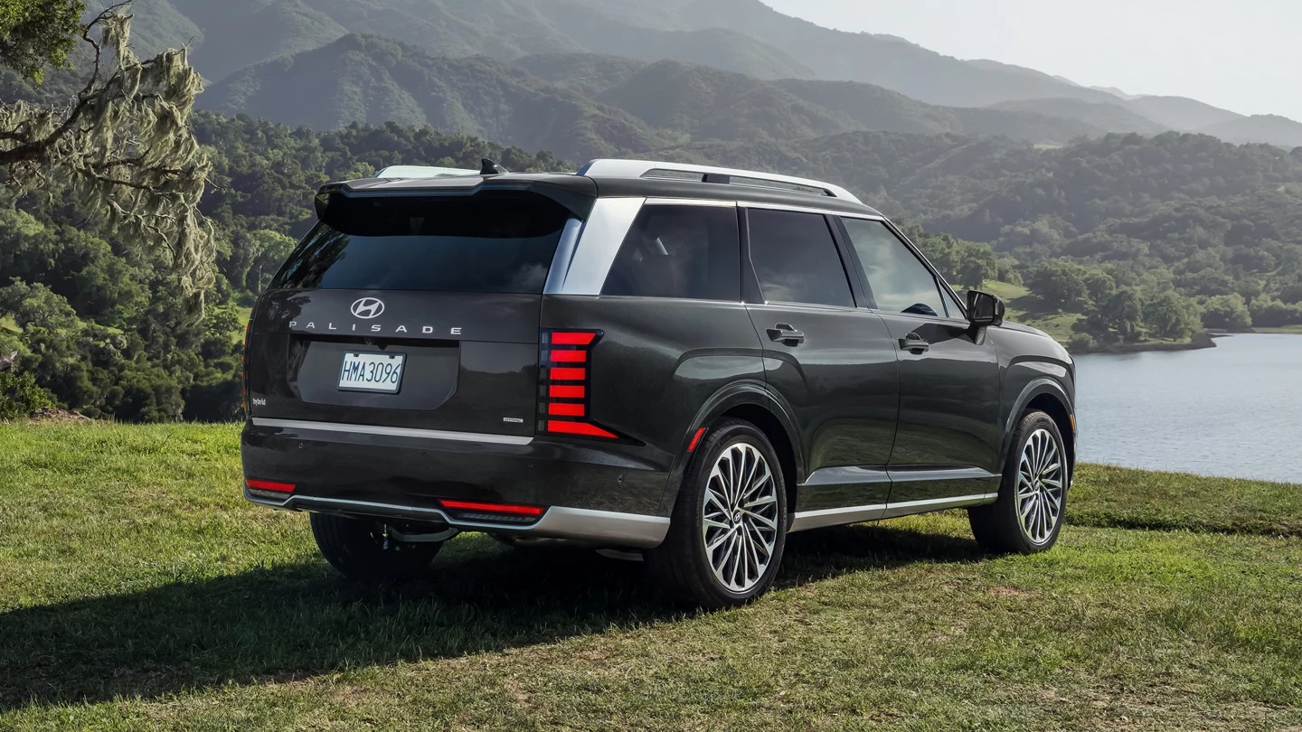 A rear view of a black Hyundai Palisade Hybrid overlooking a
lake on a grassy cliff in a mountainous region.
