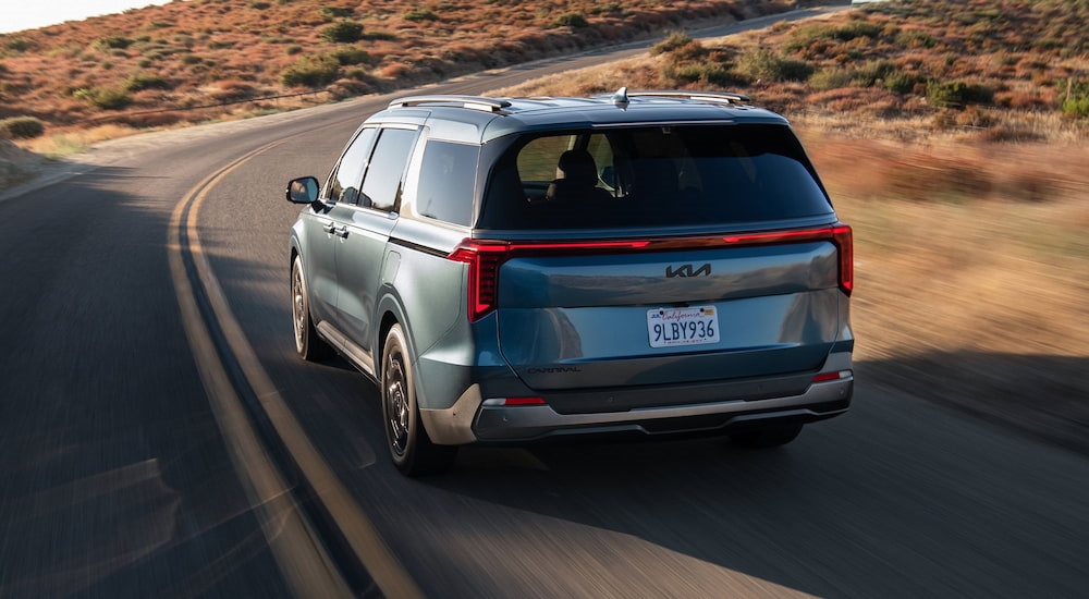 Rear of a blue 2026 Kia Carnival driving down a road.