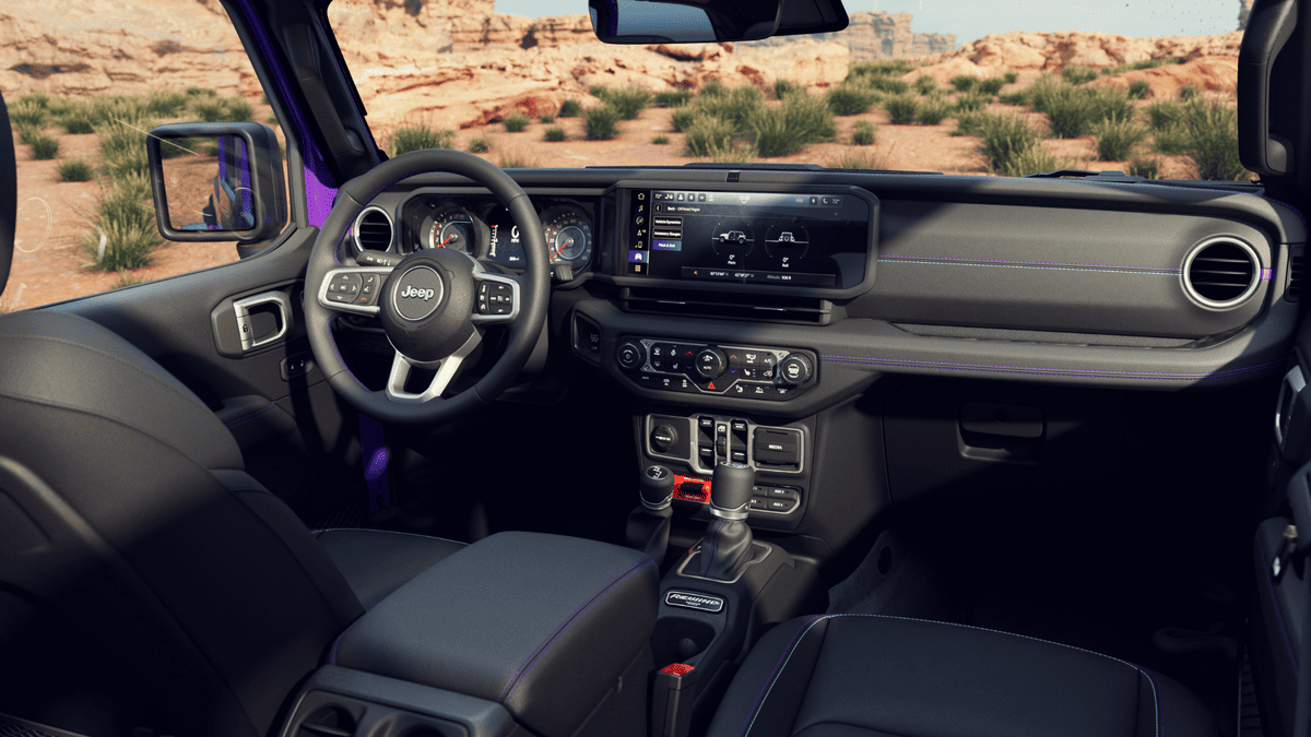 2026 Jeep Wrangler cockpit in Margate FL featuring Jeep-branded steering wheel, wide touchscreen infotainment display, manual gear shifter, and purple accent stitching with desert canyon backdrop