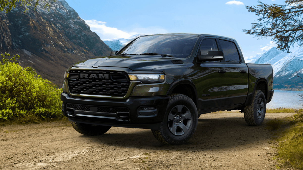 2026 Ram 1500 in Margate FL in dark olive green with black RAM grille, crew cab body style, off-road tires, and blacked-out wheels parked on a dirt trail near a mountain lake
