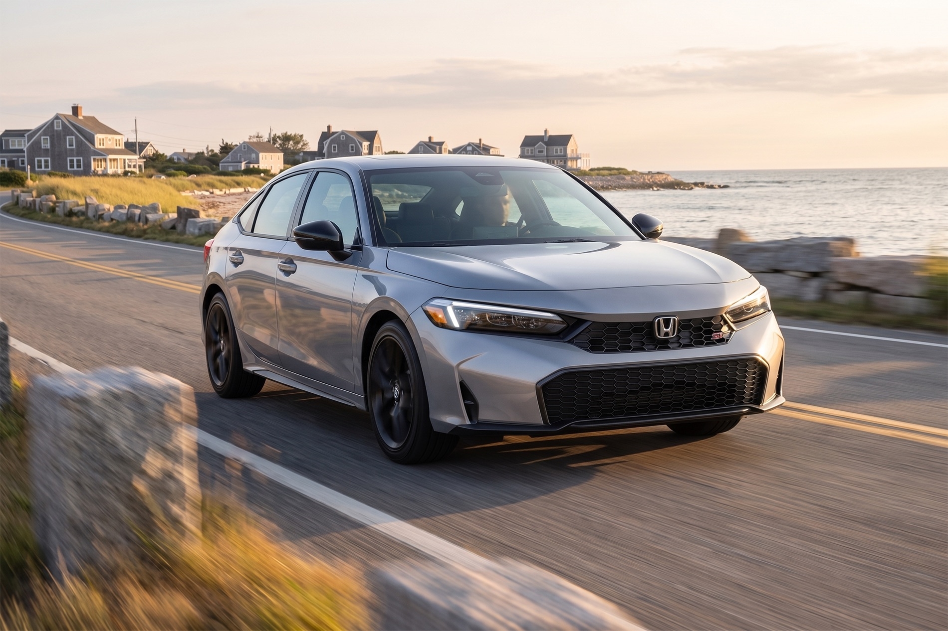 2026 Honda Civic Si driving along a coastal South Shore Massachusetts road at sunset, with ocean views, stone barriers, and seaside homes in the background under warm golden-hour light