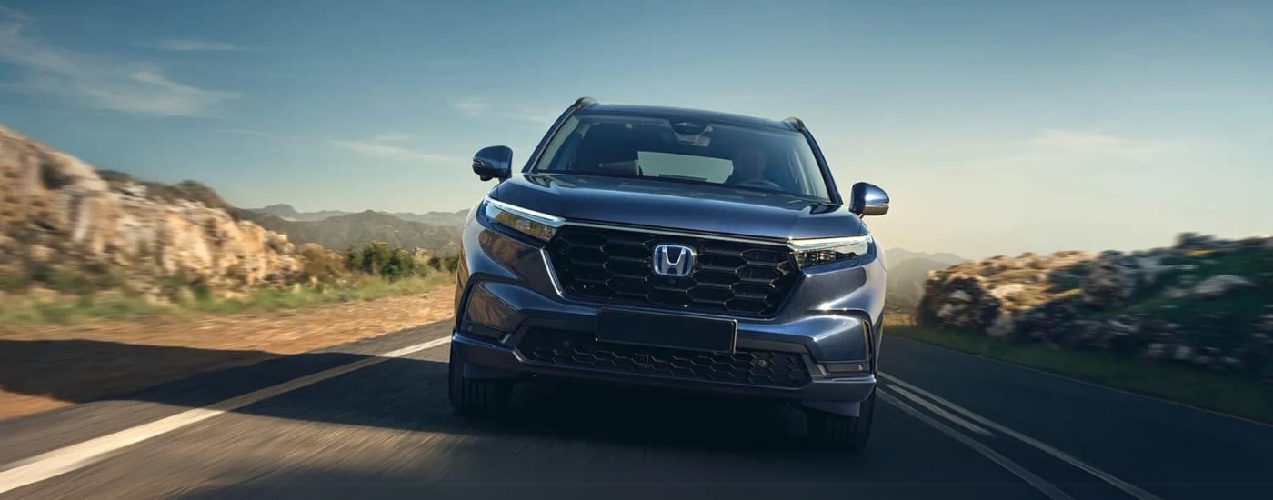 Front of a dark blue 2026 Honda CR-V Hybrid driving down a road.