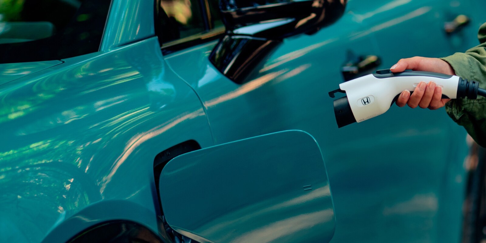 Close-up of a Honda Prologue charging outdoors with a driver holding a Honda-branded EV charging connector beside the open charging port on a blue SUV.