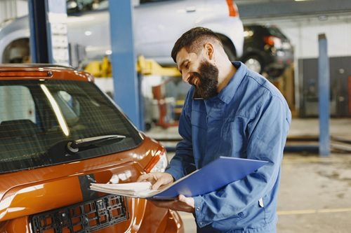 Smiling auto mechanic inspects a car in a service garage, wearing coveralls and holding documents