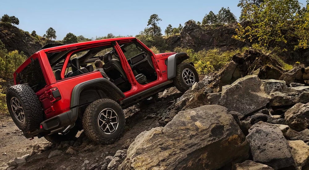Side of a red 2026 Jeep Wrangler driving off-road up a hill.