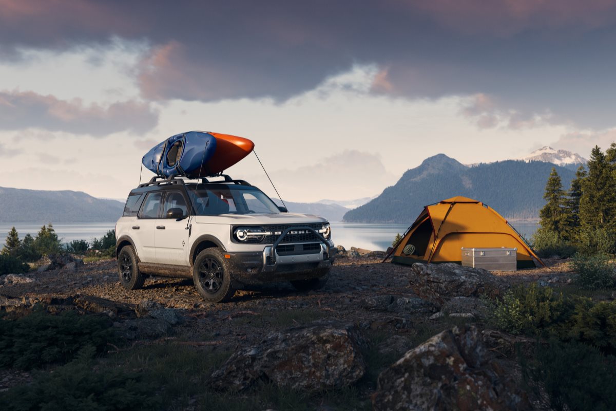 White Ford Bronco Sport parked at a lakeside campsite with a kayak mounted on the roof and a tent nearby, showcasing the SUV’s outdoor capability for camping, road trips, and summer adventures.