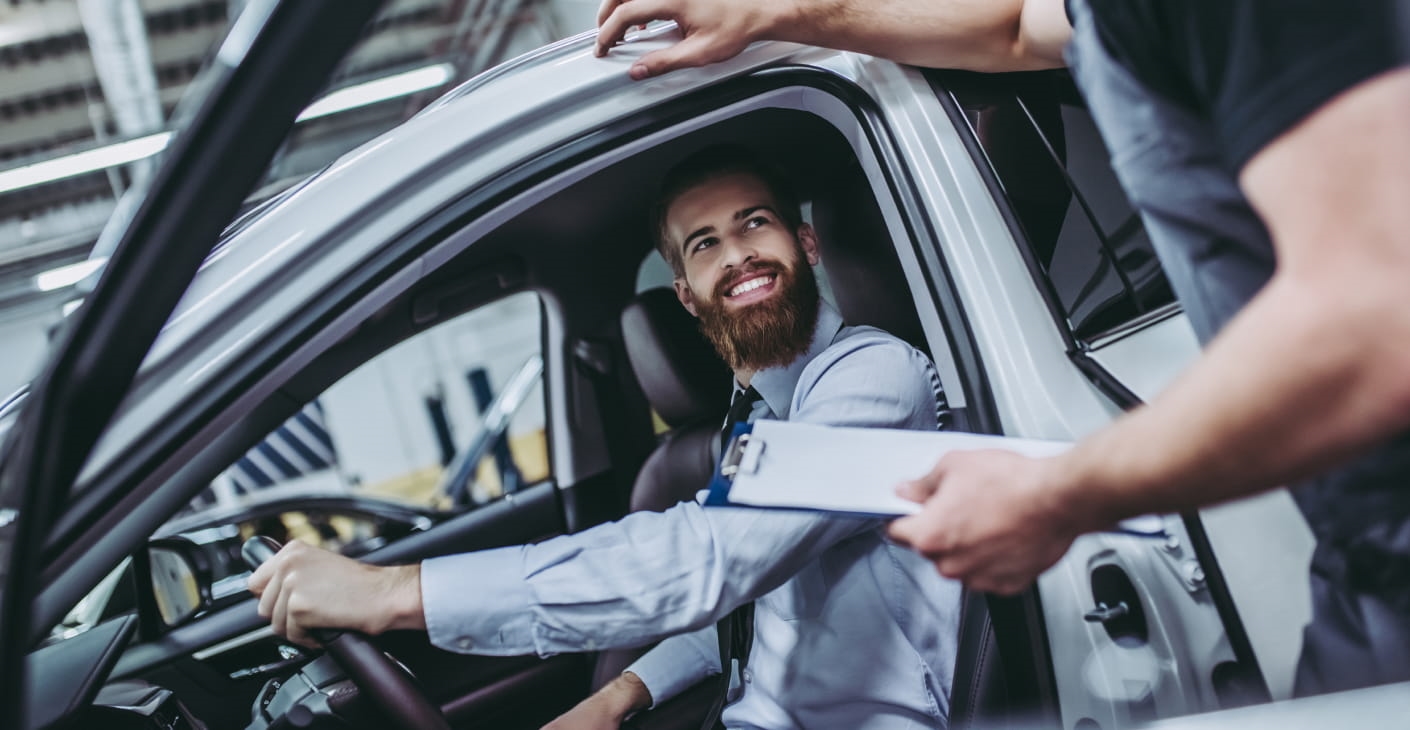 Man in a car smiles at a mechanic holding a clipboard, conveying a sense of satisfaction and professionalism in an auto service setting.
