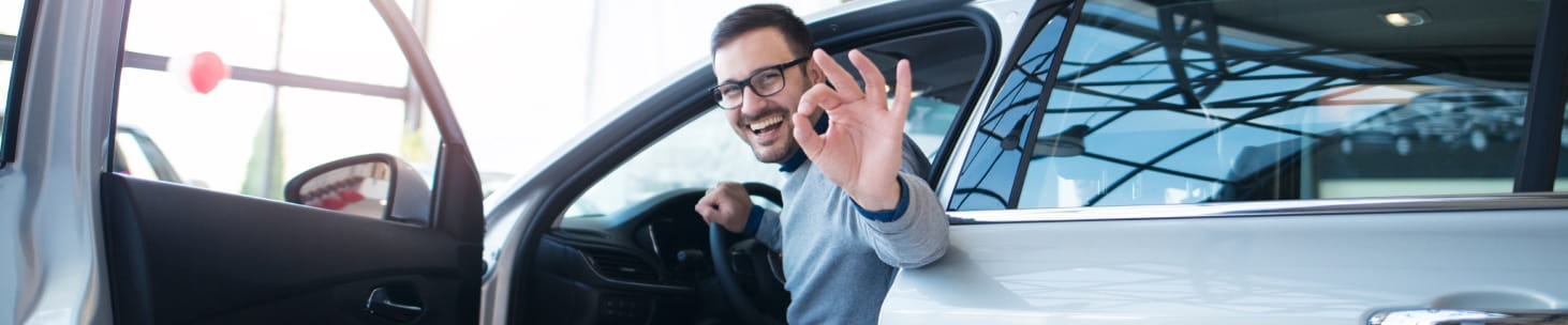 Man inside a car with door open giving a positive hand gesture in a dealership showroom.