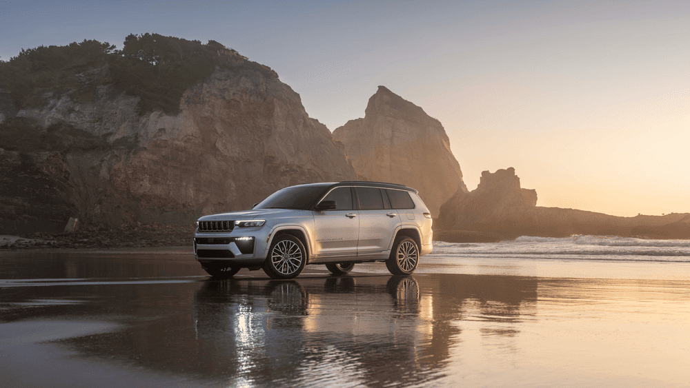 2026 Jeep Grand Cherokee L in Margate FL in silver with seven-slot grille, LED headlights, and premium alloy wheels parked on a wet beach at golden hour with coastal rock formations