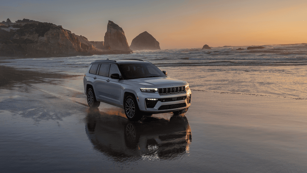 2026 Jeep Grand Cherokee L in white driving along a wet beach at sunset with LED headlights and full-width LED lightbar illuminated, showcasing smart safety technology for roads near Margate FL