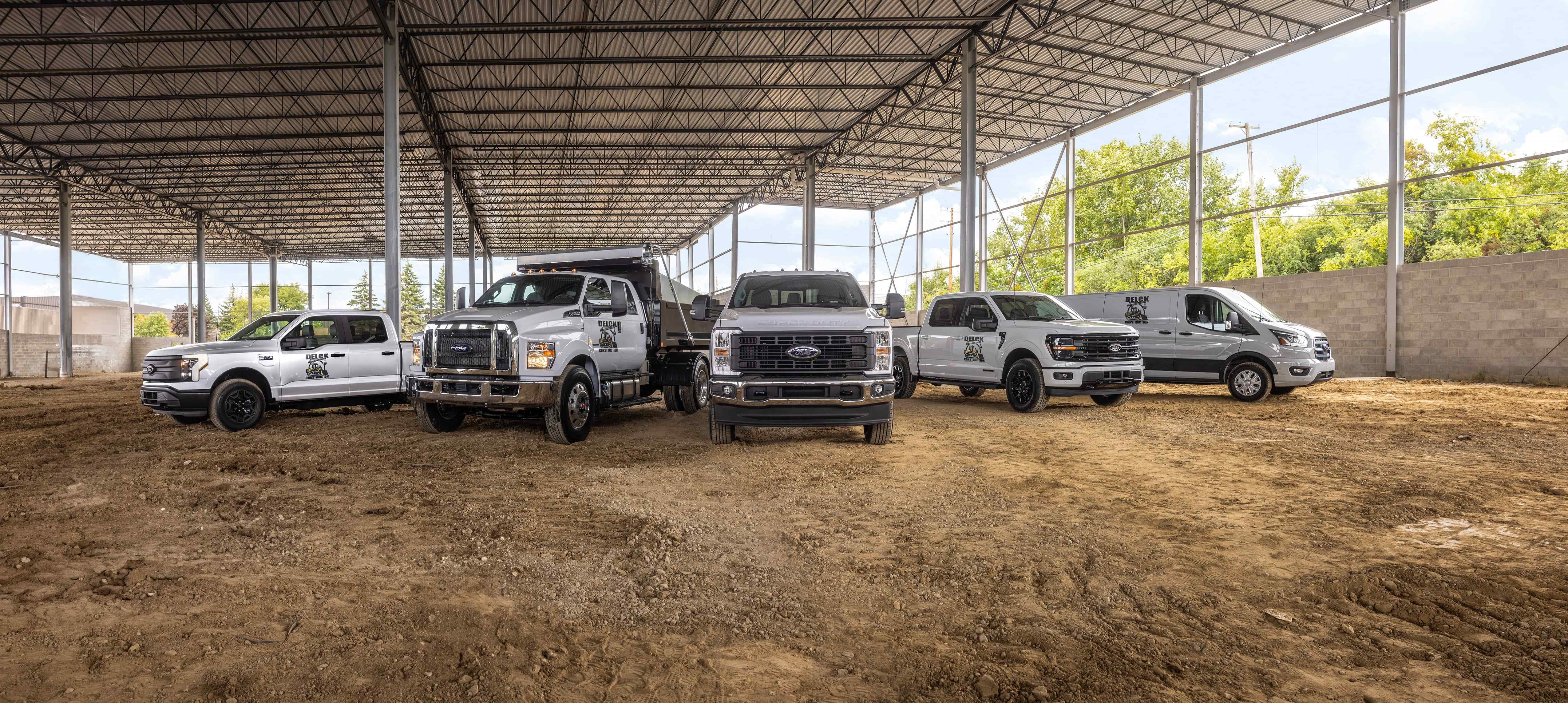 Ford commercial vehicles, including Transit vans, F-150 pickups, Super Duty trucks, and a dump truck, parked inside a large commercial building under construction.