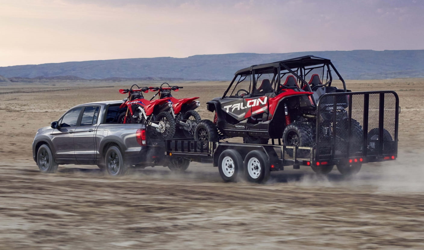 2026 Honda Ridgeline drives through a dusty desert landscape and towing a trailer with two red bikes.