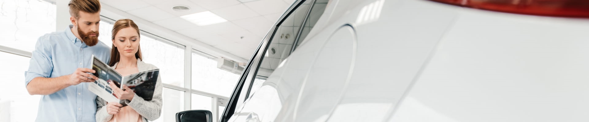 A couple examines a car brochure inside a bright showroom, standing next to a white car.