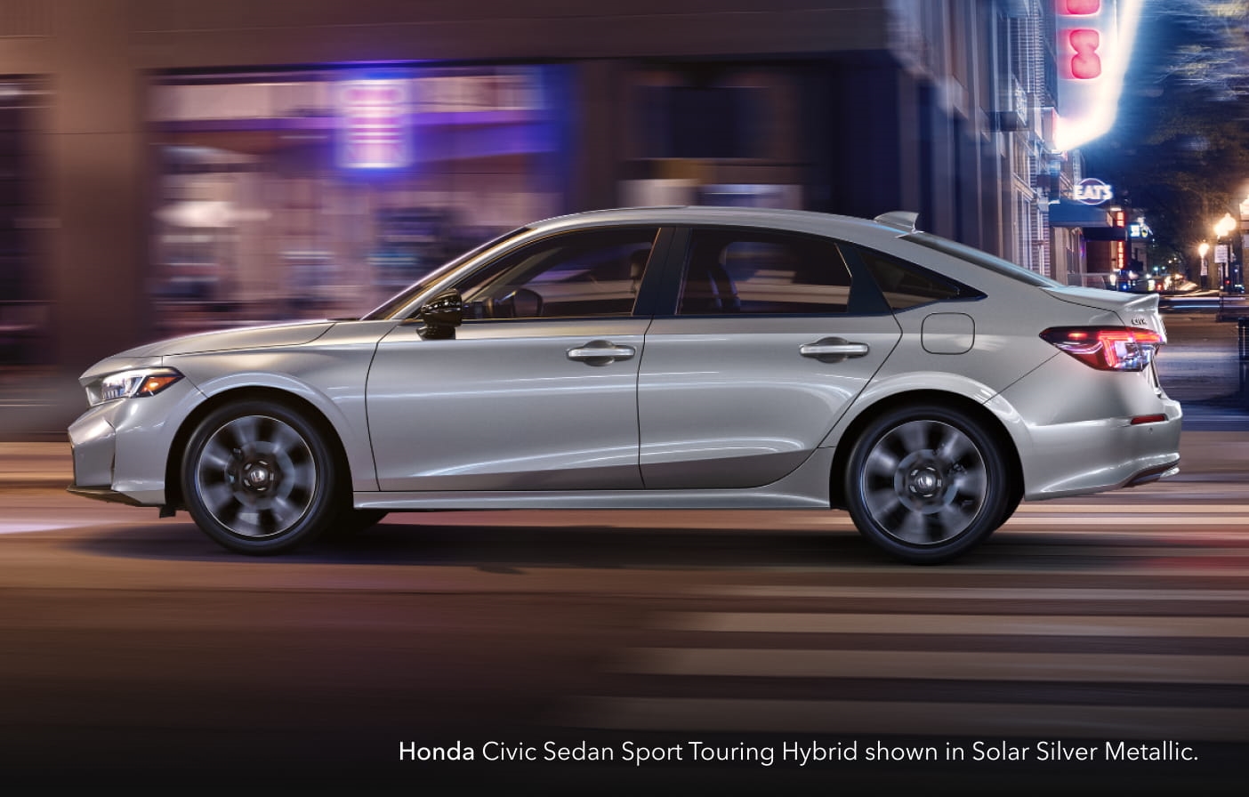 2026 Honda Civic Sedan Sport Touring Hybrid in Solar Silver Metallic drives through a vibrant city street at night.