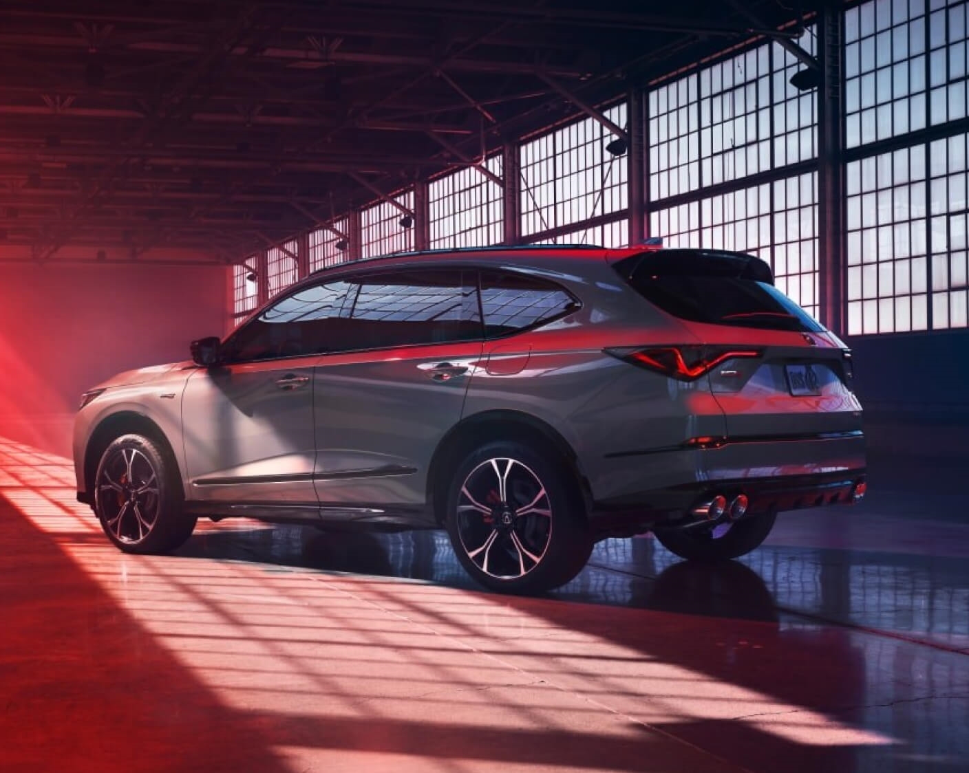 A rear three-quarter view of a gray Acura MDX parked in a dark warehouse with red lighting.
