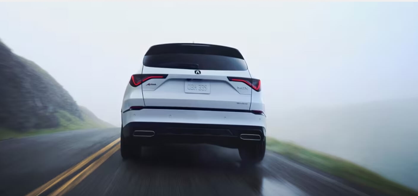 A rear view of a white Acura MDX driving on a foggy cliffside road.