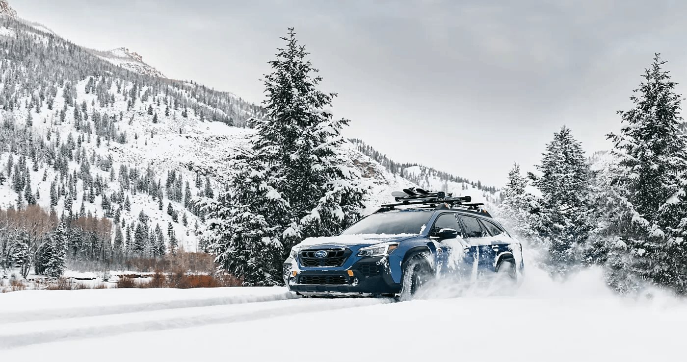 A blue Subaru Outback with skis on the roof rack driving through fresh snow, pine forest and snowy mountains behind.