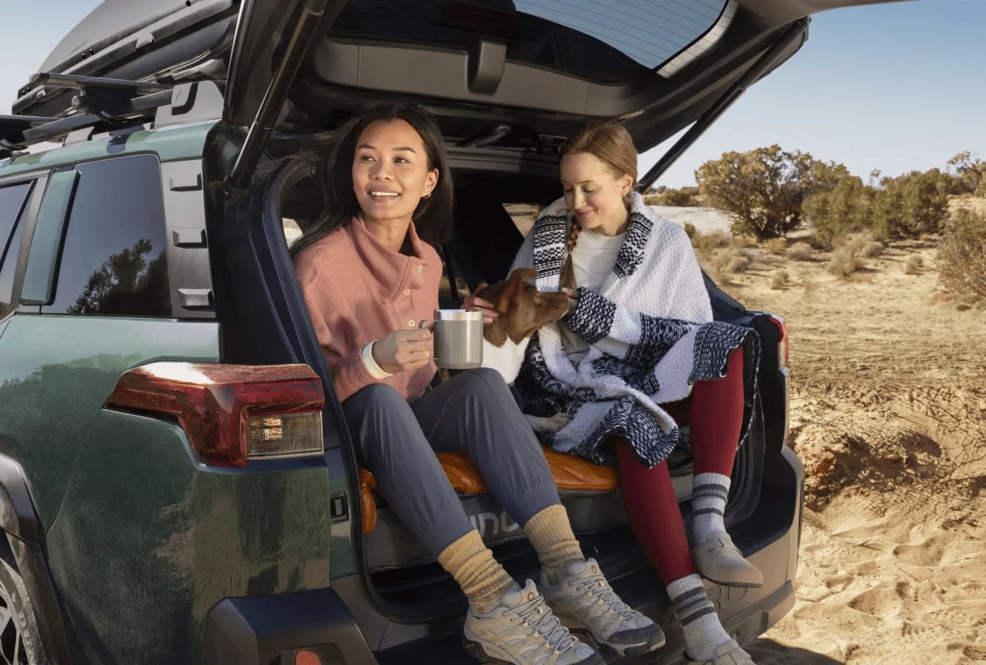 Two friends sitting in the open cargo area of a Subaru Outback with coffee and a dog, desert behind them.