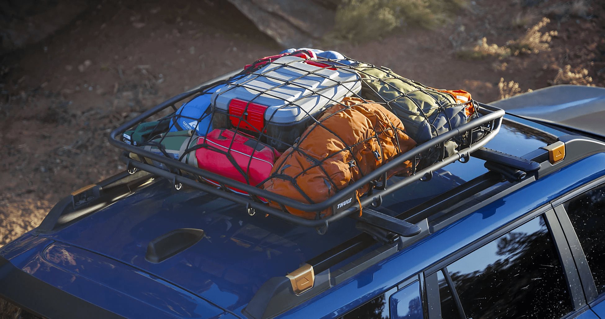 A roof basket on a Subaru Outback packed with camping gear, coolers, and bags on a dirt trail.