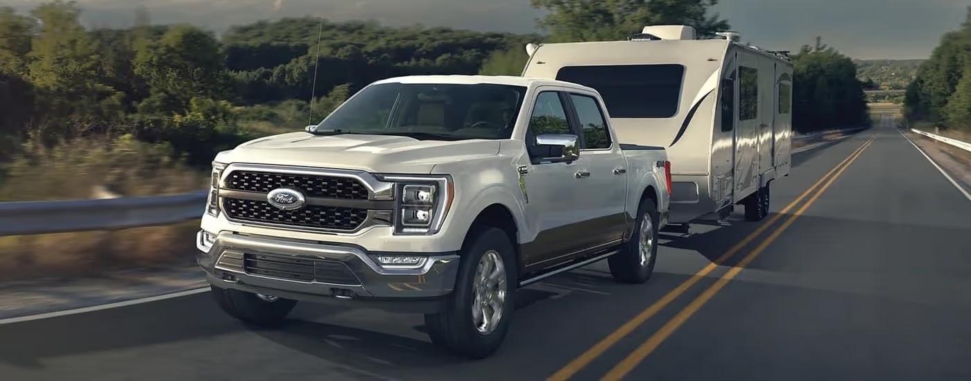 A white 2023 Ford F-150 is shown towing a camper trailer on an open road.