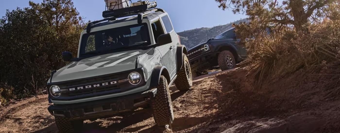 A grey and a blue 2023 Ford Bronco are shown driving on a trail.