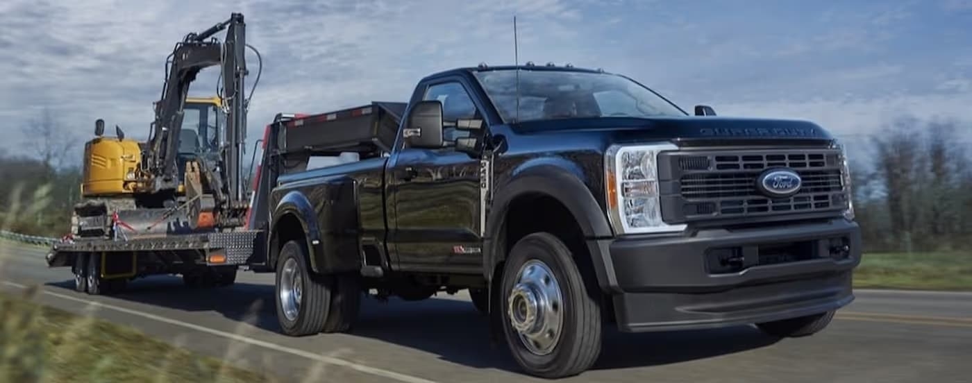 A black 2025 Ford F-450 towing heavy machinery on a highway.