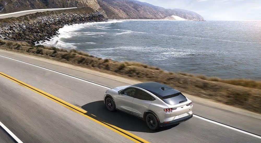 A white 2024 Ford Mustang Mach-E is shown driving near a coast.