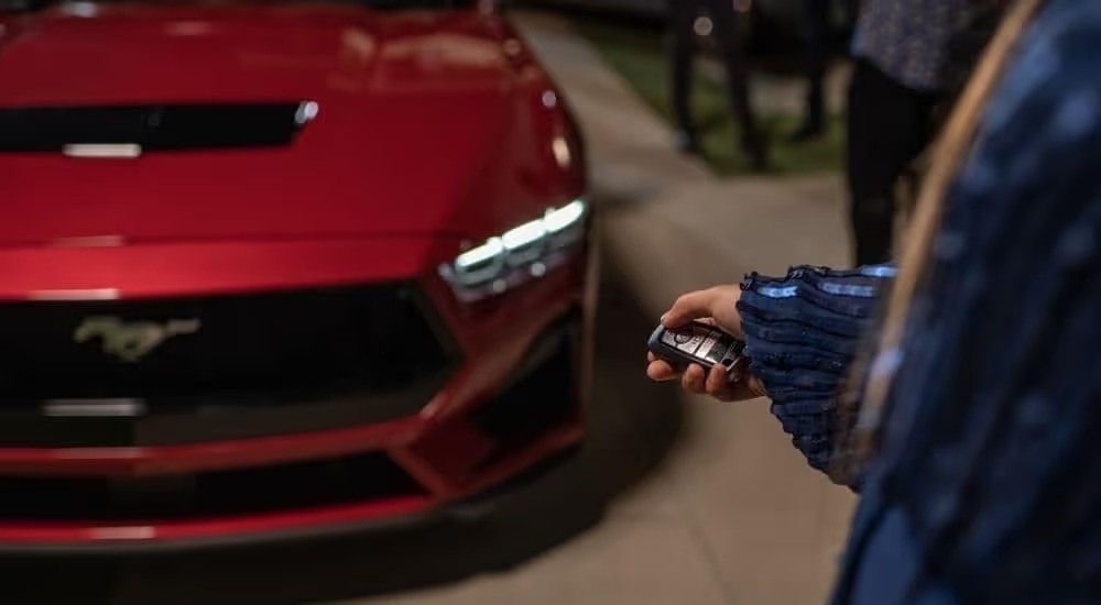 A person is shown pointing a key fob at the hood of a red 2024 Ford Mustang.