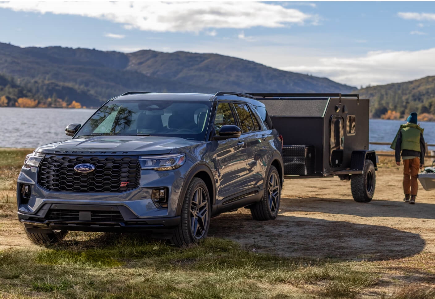 Three-quarter front view of a Ford Explorer ST in Vapor Blue towing a trailer on a shore with a walking family.