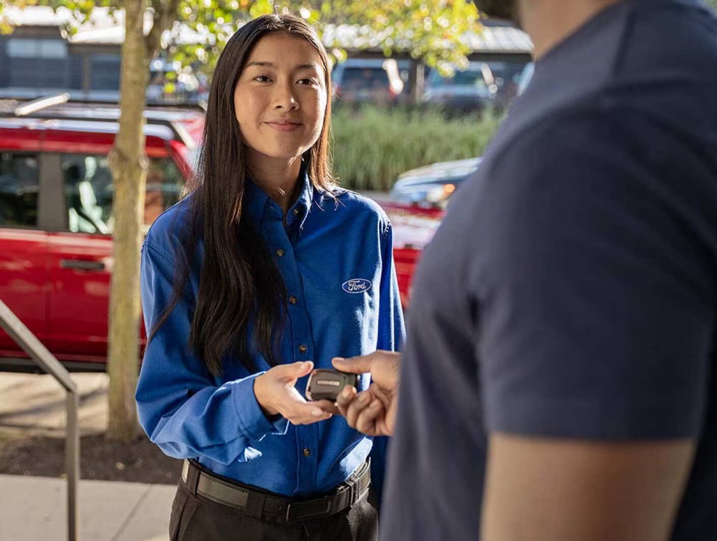 A Ford service advisor handing a vehicle key fob to a customer outside of a dealership lot.