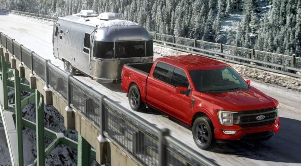 A red 2024 Ford F-150 shown pulling a camper over a snowy bridge.